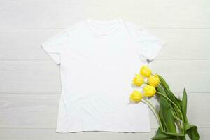 blanco camiseta Bosquejo. modelo blanco camisa parte superior vista. blanco de madera antecedentes. madre mujer día día festivo. amarillo tulipanes mujer camiseta con cumpleaños ramo de flores flores primavera mirar. hembra accesorios foto