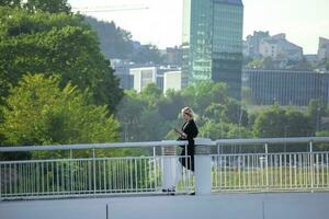 Vilna, Lituania 03 06 2022 joven mujer caminando en un blanco puente con célula teléfono en un antecedentes con verde arboles y rascacielos foto