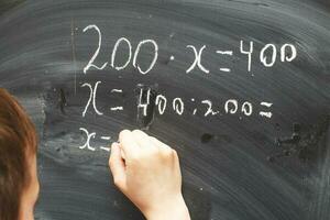 Boy standing back in front of school blackboard and writing. Schoolboy solves math example at the chalkkboard photo