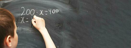 Boy standing back in front of school blackboard and writing. Schoolboy solves math example at the chalkkboard photo