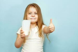 pequeño hermosa niña participación Leche en vaso en un azul antecedentes. niño bebidas yogur foto