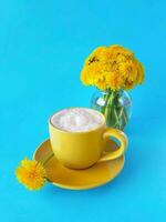 Coffee topped with whipped cream in yellow cup and one dandelion on saucer. Bouquet of fresh dandelions in little glass vase on blue backgroung. Hello summer concept. Vertical. photo