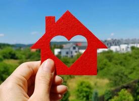 Hand holding red felt house on blurred background of cottage village in summer sunny day. Heart-shaped hole is cut instead of a window. Buy or build your dream home, real estate agency concept. photo