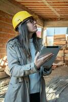 Vertical portrait of female architect with yellow hard hat standing looking at construction site with her tablet in hand. photo