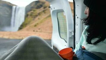 Tourist woman sit hold cup enjoy nature views with open door inside rented campervan. Skogafoss waterfall, Iceland. Travel solo sightseeing Iceland. Stay official campsite video