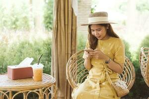 Asian woman with beautiful smile reading good news on mobile phone during rest in coffee shop, happy female watching her photos on cell phone while relaxing in cafe during free time