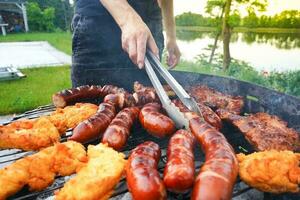 Man turning the sausage on the bbq with kitchen tongs. Person grilling in the garden. Barbeque with sausages and fried fish on the grill. photo