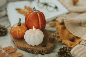 Cozy autumn decor - burning candle shape of pumpkin and orange decor pumpkins on wooden board on bed with open book, warm sweater, autumn leaves photo
