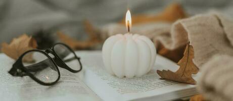 White burning candle shape of pumpkin on open book, eyeglasses, cozy sweater and autumn leaves on bed. Long banner for design, web site photo