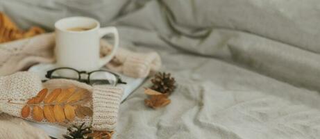 Cozy home atmosphere. Warm beige sweater, mug with tea or coffee, book and eyeglasses on gray bed in bedroom. Hygge concept. Long banner with copy space for design, web site photo