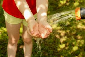 un niño pone su manos debajo agua fluido desde un riego manguera en un calentar verano día foto