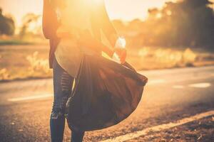 mujer son coleccionar basura en el la carretera con el fondo de el puesta de sol. limpiar arriba a reducir global calentamiento foto