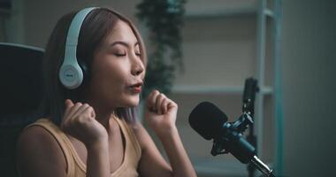 antal fot av ung asiatisk kvinna influencer bär hörlurar talande in i en mikrofon medan inspelning en radio visa i en levande rum Hem studio. innehåll skapare och influencer marknadsföring begrepp. video