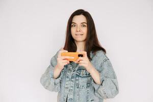 indeciso y inseguro. joven mujer en mezclilla chaqueta jugando teléfono inteligente juego, aislado en gris antecedentes. enfatizando incertidumbre y duda foto