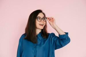 Smiling woman touching glasses, pondering and looking for ideas in empty space. Isolated portrait on pink background photo