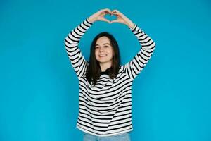 Joyful Woman in Striped Sweater Expressing Love and Happiness - Happy Girl Showing Heart Gesture with Arms, Smiling - Isolated Blue Background with Copy Space - Perfect for Love, Happiness Concepts photo