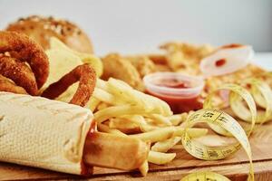 Unhealthy and junk food. Different types of fastfood and snacks on the table with measuring tape photo