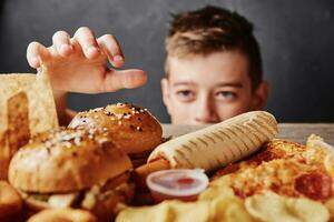 hambriento chico mira a sabroso comida y tomar un hamburguesa desde mesa. insalubre comida concepto foto