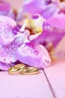 Golden wedding rings with purple orchids on pink wooden table. Vertical. Wedding ceremony, honeymoon, marriage proposal, Valentine's day in hot countries. Copy space photo