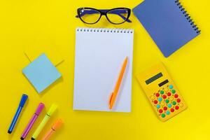 cuaderno con bolígrafo, marcadores y pegatinas con calculadora y lentes en amarillo antecedentes. espalda a escuela, negocio reunión. Copiar espacio foto