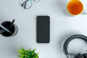 Phone on work desk. Top view, flat lay composition. Surrounded by coffee mug, headset, plant, glasses and pens photo