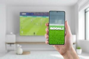 Goal on the phone display. The concept of tracking sports results online. Living room with a TV and soccer game in background photo