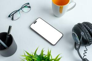 Phone mockup with isolated screen for app presentation placed on office desk, surrounded by coffee mug, glasses, pens, plant and headset. photo