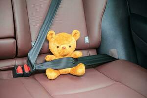 Yellow bear doll fastening seat belt during sitting inside a car and driving on the road. Car Seat, safety, trip, journey and transport concept photo