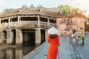 Woman traveler wearing Ao Dai Vietnamese dress sightseeing at Japanese covered bridge in Hoi An town, Vietnam. landmark and popular for tourist attractions. Vietnam and Southeast Asia travel concept photo