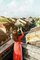 happy woman wearing Ao Dai Vietnamese dress, traveler taking photo by mobile phone on rooftop at Hoi An ancient town in Vietnam. Vietnam and Southeast travel concept