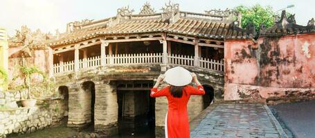 Woman traveler wearing Ao Dai Vietnamese dress sightseeing at Japanese covered bridge in Hoi An town, Vietnam. landmark and popular for tourist attractions. Vietnam and Southeast Asia travel concept photo