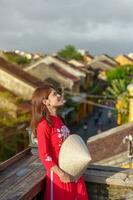 happy woman wearing Ao Dai Vietnamese dress, traveler sightseeing view at rooftop at Hoi An ancient town in Vietnam. landmark and popular for tourist attractions. Vietnam and Southeast travel concept photo