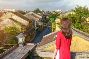 happy woman wearing Ao Dai Vietnamese dress, traveler sightseeing view at rooftop at Hoi An ancient town in Vietnam. landmark and popular for tourist attractions. Vietnam and Southeast travel concept photo