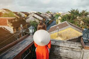 happy woman wearing Ao Dai Vietnamese dress, traveler sightseeing view at rooftop at Hoi An ancient town in Vietnam. landmark and popular for tourist attractions. Vietnam and Southeast travel concept photo