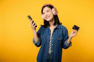 retrato joven asiático mujer con contento éxito sonrisa vistiendo mezclilla ropa y auricular participación teléfono inteligente y crédito tarjeta. compras en línea móvil teléfono entretenimiento estilo de vida concepto. foto