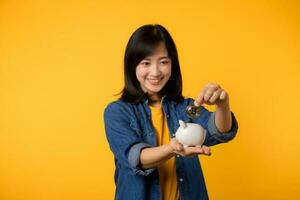 Happy young asian woman wearing yellow t-shirt denim shirt saving digital crypto currency to piggy bank isolated on yellow background. Digital money saving investment concept. photo