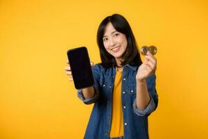 Happy young asian woman wearing yellow t-shirt denim shirt holding digital crypto currency coin and smartphone isolated on yellow background. Digital currency financial concept. photo