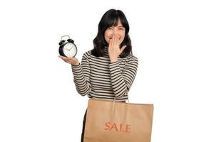 Portrait of excited young Asian woman with sweater shirt holding alarm clock and shopping paper bag isolated on white background. last deals shocked price shopping concept photo