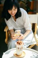 Smiling asian woman taking photo of ice coffee with smartphone camera in coffee cafe.