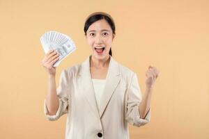 retrato de contento exitoso confidente joven asiático negocio mujer vistiendo blanco chaqueta participación efectivo dinero dolares en pie terminado beige antecedentes. millonario negocio, compras concepto. foto