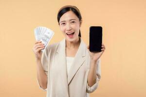 portrait of happy successful confident young asian business woman wearing white jacket holding cash money dollars and credit card standing over beige background. millionaire business, shopping concept photo