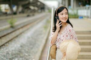 Asian young woman traveler with weaving hat using a mobile phone beside railway train station in Bangkok. Journey trip lifestyle, world travel explorer or Asia summer tourism concept. photo