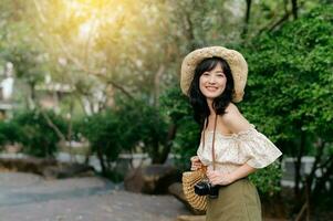 Portrait of asian young woman traveler with weaving hat and basket and a camera on green public park nature background. Journey trip lifestyle, world travel explorer or Asia summer tourism concept. photo