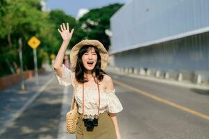 Portrait of asian young woman traveler with weaving hat and basket and a camera waving hand to friend by the street. Journey trip lifestyle, world travel explorer or Asia summer tourism concept. photo