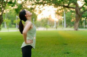 Female jogger. Fit young Asian woman with green sportswear breathing fresh air in park before running and enjoying a healthy outdoor. Fitness runner girl in public park. Wellness being concept photo