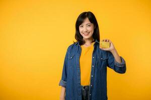 retrato de joven asiático mujer vistiendo amarillo camiseta y mezclilla chaqueta con contento sonrisa participación un crédito tarjeta aislado en amarillo antecedentes. pago compras en línea concepto. foto