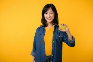 retrato de joven asiático mujer vistiendo amarillo camiseta y mezclilla chaqueta con contento sonrisa participación un crédito tarjeta aislado en amarillo antecedentes. pago compras en línea concepto. foto