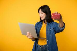 contento joven mujer de asiático etnia vestir amarillo camiseta mezclilla camisa utilizando ordenador portátil ordenador personal computadora sostener crédito banco tarjeta compras en línea orden entrega aislado en llanura amarillo antecedentes. foto