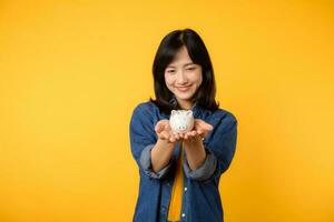 portrait cheerful young asian woman wearing yellow t-shirt denim shirt with happy smile holding piggybank save investment money isolated on yellow background. economy finance business wealth concept. photo