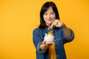 Happy young asian woman wearing yellow t-shirt denim shirt saving digital crypto currency to piggy bank isolated on yellow background. Digital money saving investment concept. photo